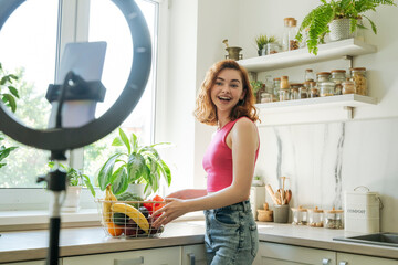 Happy influencer with basket of fruits and vegetables standing by kitchen counter at home