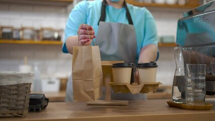 Close-up takeaway coffee cups and paper bag in female Caucasian hands in cafe. Unrecognizable young barista stretching drink and dessert standing in coffee house indoors
