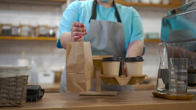 Close-up takeaway coffee cups and paper bag in female Caucasian hands in cafe. Unrecognizable young barista stretching drink and dessert standing in coffee house indoors