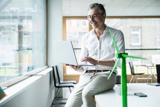 Smiling businessman sitting on desk using laptop by wind turbine model in office