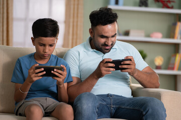 Indian father with son busy playing video game on mobile phone while sitting on sofa at home -...