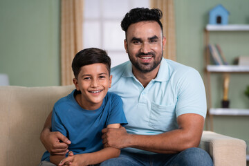 Happy smiling indian father with son by looking at camera while sitting on sofa at home - concept...