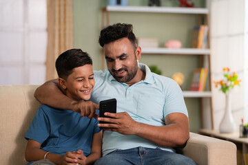 Happy smiling indian father showing mobile phone to son while sitting on sofa at home - concept of...