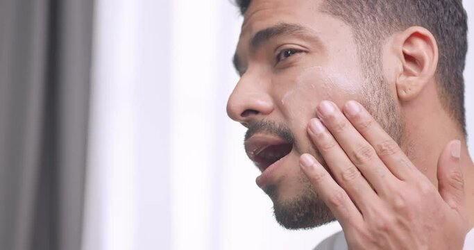 Handsome young man applying cream at his face while standing in front of mirror in bathroom at home.