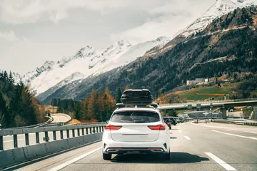 Abwaschbare Fototapete Alpen Rooftop cargo carrier bag. Rear view of a car with a roof box. Alpine highway. Black Roof Box on a Sporty White Wagon Family Car. Removable black car trunk for luggage on the roof of a car.