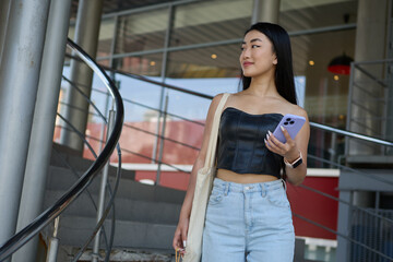 Fashionable young Asian woman walking in a city mall with a smart phone in hand. Cheerful Vietnamese female person went shopping with a tote bag