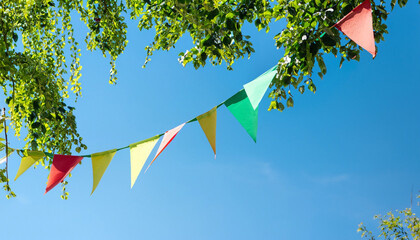 colorful pennant string decoration in green tree foliage on blue sky, summer party background...