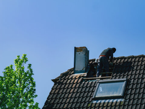 A roofer stands atop a tall house, demolishing the chimney as clear blue sky stretches above them. The buildings architecture and tiled roofs remain majestic beneath their feet.
