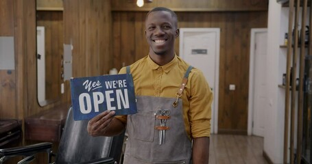 Joyful barber in apron holding welcoming Yes are open banner standing in modern salon smiling. Successful barbershop start-up and people concept.