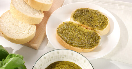 bread with pesto on a white plate. basil and slices of bread on a white tablecloth. traditional italian green sauce. angle view. sunny day.