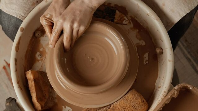 Potter making ceramic bowl or plate on pottery wheel. Top view. Potter shapes clay product. Two hands create bowl. Close-up in 4K, UHD
