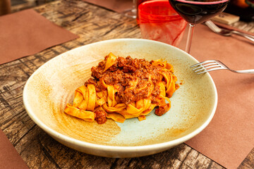 Italian Dinner in restaurant. Tagliatelle all ragu, traditional type of pasta, egg noodles served with classic  meat sauce or Bolognese sauce, brown wooden table. Red wine glass. Lifestyle photo.