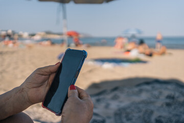 Hands of retired senior woman holding her mobile on vacation. Close-up image of an elderly lady in the sand on the beach looking at her mobile phone.