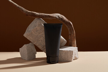 Minimalist scene for advertising with a black cosmetic bottle unbranded on brown background with blocks of stone and dry twig. Mockup for facial cleanser, lotion or moisturizer. Space for design.