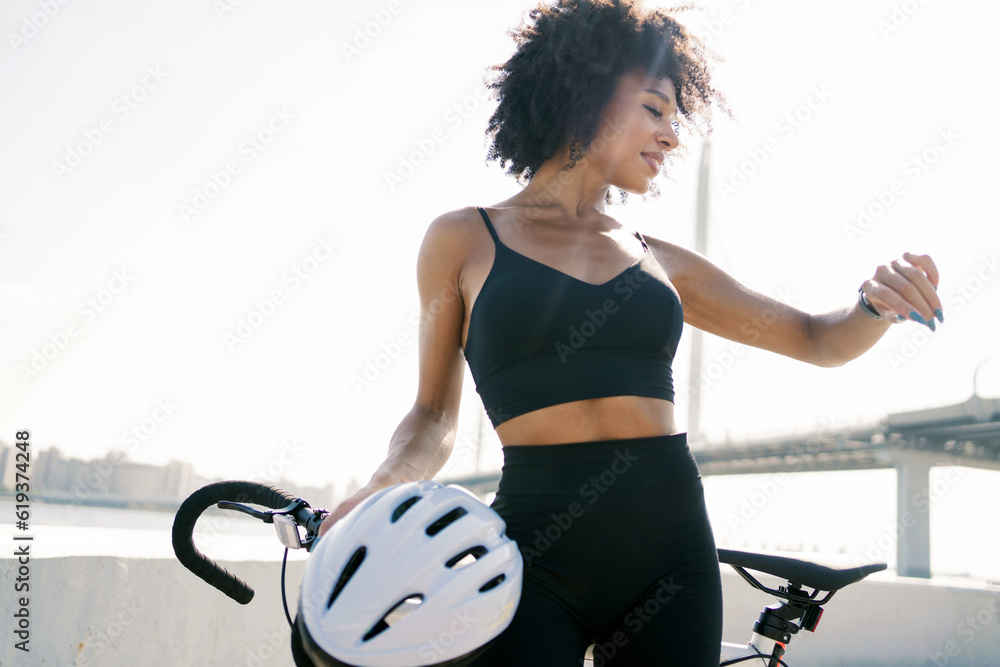 Wall mural A sporty woman looks at a fitness watch and uses an application for cycling workouts outdoors. He holds a bicycle helmet in his hand.