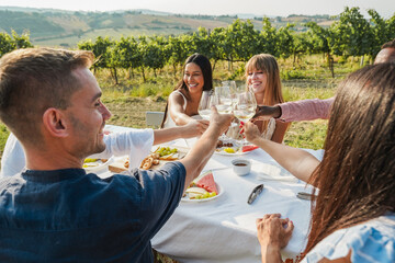 Happy multiracial friends cheering at summer dinner with vineyard in background - Adult people enjoying white wine glass at countryside resort - Focus on closeup hands