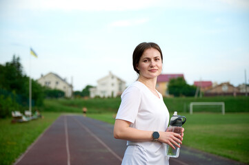 Portrait of the slim fit young girl with bottle of water. Sporty female standing near the stadium on the jogging track.