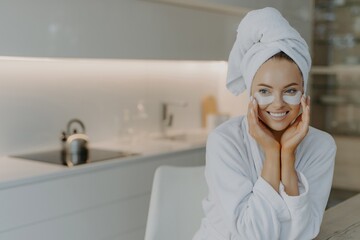 Woman with healthy skin applies recovery pads under eyes, enjoys beauty treatment. Soft bathrobe, wrapped towel. Home interior. - obrazy, fototapety, plakaty