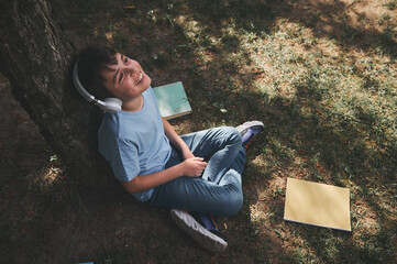 Top view Caucasian schoolboy in headphones, relaxing sitting under the tree in the park, listening to music after class