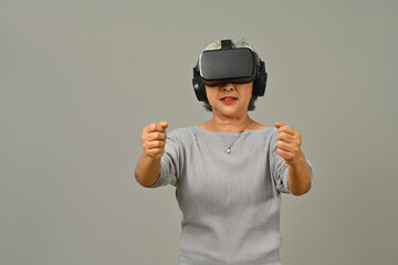 Smiling middle age woman with virtual reality glasses simulating of driving car in VR glasses isolated on gray background