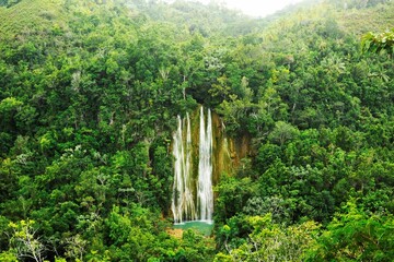 Majestic waterfall cascading over lush green hills in a tropical environment, framed by lush trees. - obrazy, fototapety, plakaty