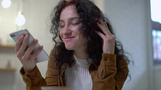 Young woman using voice control turning on playlist in smartphone app. Portrait of satisfied Caucasian lady enjoying music in headphones sitting in cafe indoors