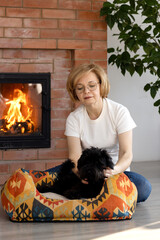 A beautiful middle-aged woman sitting by the fireplace next to her dog on a couch.