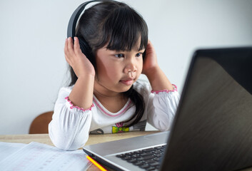 5-6 years old Asian girl wearing white shirt is studying online via computer using bluetooth headphones at home, distance education concept