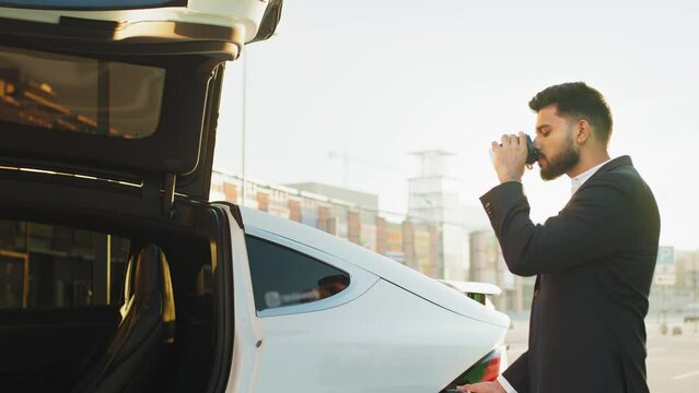 Arabian businessman in suit inserting power cord into white electric car while standing outdoors with takeaway coffee in one hand. Zero emission vehicle. Indian man at station charging electro car.