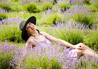 beautiful brunette smiling woman in violet dress  and black hat sitting in the blooming field of...