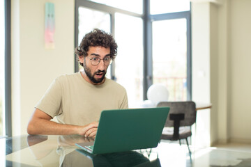 young adult bearded man with a laptop looking very shocked or surprised, staring with open mouth saying wow