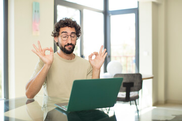 young adult bearded man with a laptop looking concentrated and meditating, feeling satisfied and...