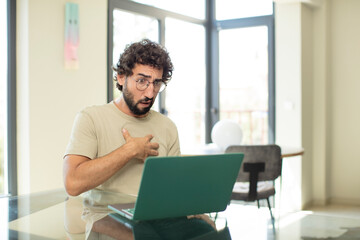 young adult bearded man with a laptop feeling shocked, astonished and surprised, with hand on chest...