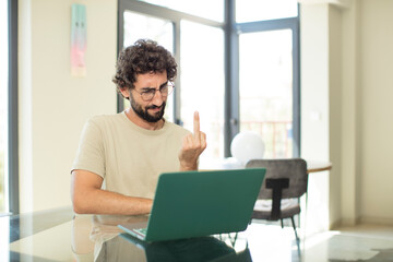 young adult bearded man with a laptop feeling angry, annoyed, rebellious and aggressive, flipping the middle finger, fighting back