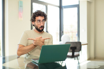 young adult bearded man with a laptop looking puzzled and confused, insecure and pointing in opposite directions with doubts - obrazy, fototapety, plakaty