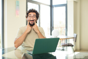 young adult bearded man with a laptop smiling and pointing to own fake smile