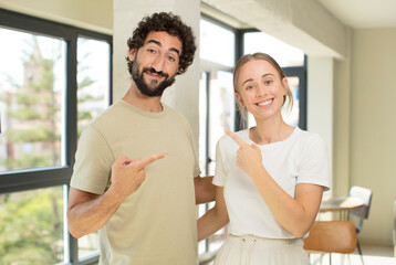 Obraz premium young adult couple smiling cheerfully, feeling happy and pointing to the side and upwards, showing object in copy space