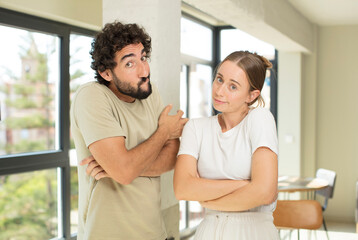 young adult couple shrugging, feeling confused and uncertain, doubting with arms crossed and puzzled look