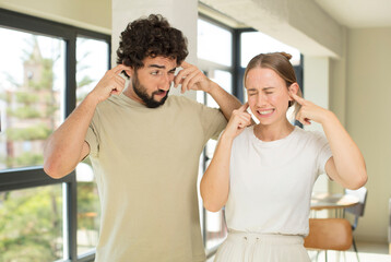 young adult couple looking angry, stressed and annoyed, covering both ears to a deafening noise, sound or loud music