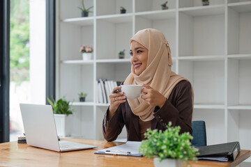 Asian Muslim business woman holding a coffee mug sitting in the office looking out the window. business people, diversity and office concept