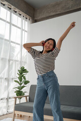 Attractive happy young Asian woman listening music with headphones from music application dance raise up hand near grey sofa couch stay at home rest relax spend free spare time in living room indoor
