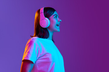 Side view portrait of happy, smiling, positive young girl listening to music in headphones against gradient purple background in neon light. Concept of emotions, youth, feelings, fashion, lifestyle