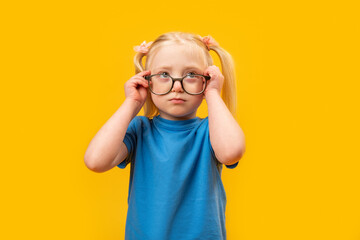 Elementary schoolgirl with two ponytails wears blue T-shirt and glasses looks up. Studio portrait of child, yellow background