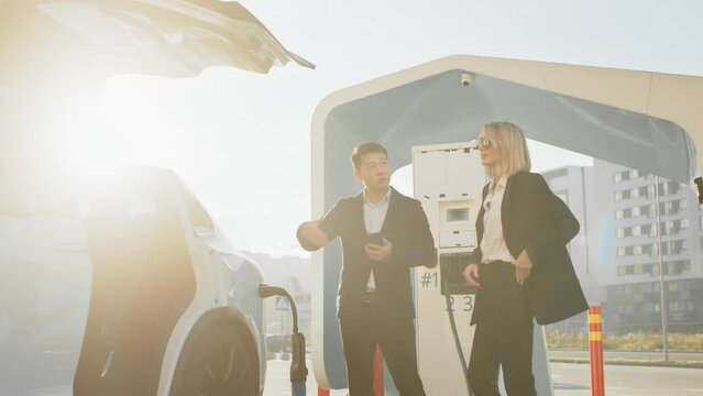 Stylish caucasian woman talking with asian car dealer while buying new electric vehicle. Multiracial people standing near moden auto on charging station. Transport and purchase concept. Electric car.
