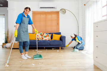 Two Asian young professional cleaning service women worker team working in the house. Girls housekeeper sweeps broomsticks on the wooden floor with another one cleaning under the sofa. Cleaner.