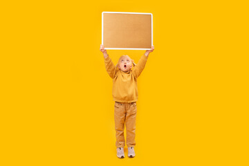 Little surprised girl in yellow suit holds cork board high above her head. Copy space. Isolated on yellow background.