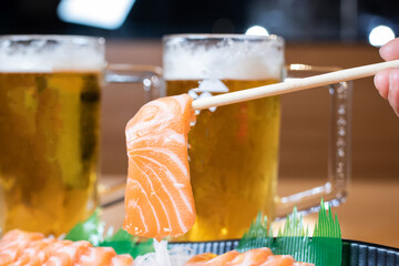 Hand holding chopsticks holding salmon sashimi served with two glasses of draft beer.