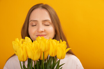 Fototapeta premium Young blonde woman enjoys scent of flowers. Girl holds bouquet of yellow tulip flowers on yellow background. Close up. Copy space.
