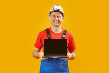 Middle aged man architect engineer wears safety helmet which hand holding laptop with empty screen on yellow studio background. Copy space, mock up.