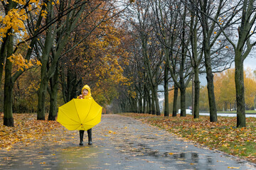 Child walks in an autumn park in rain with an umbrella in his hands. Yellow umbrella and raincoat